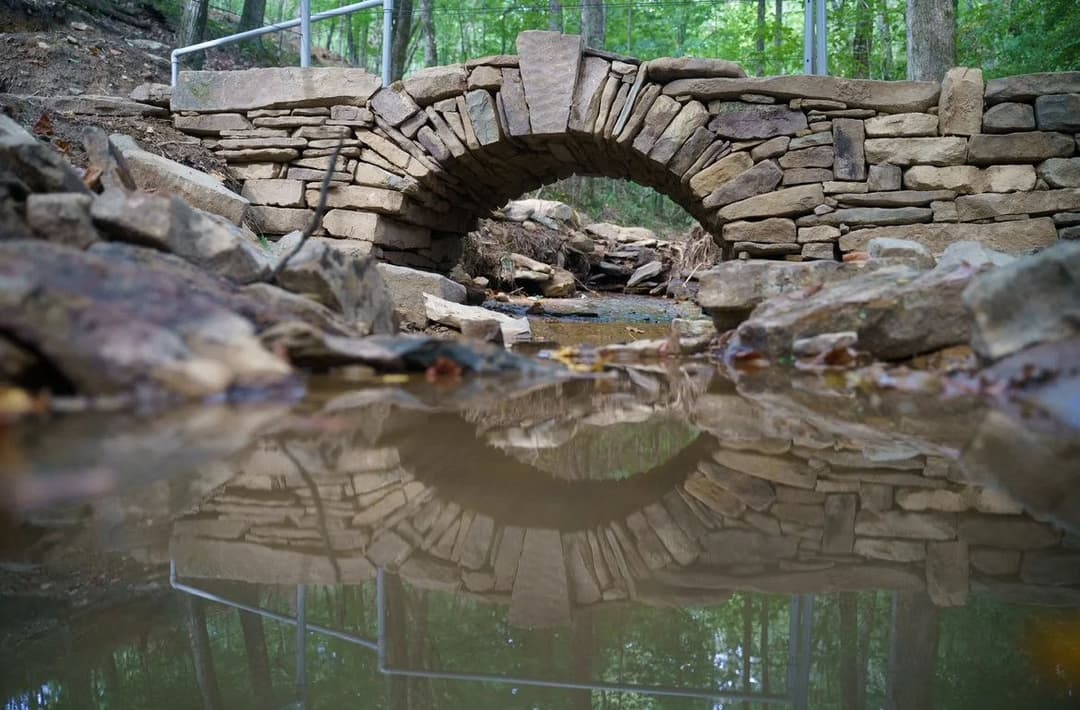 Stone arch bridge reflected in water, surrounded by lush green forest and rocky terrain.
