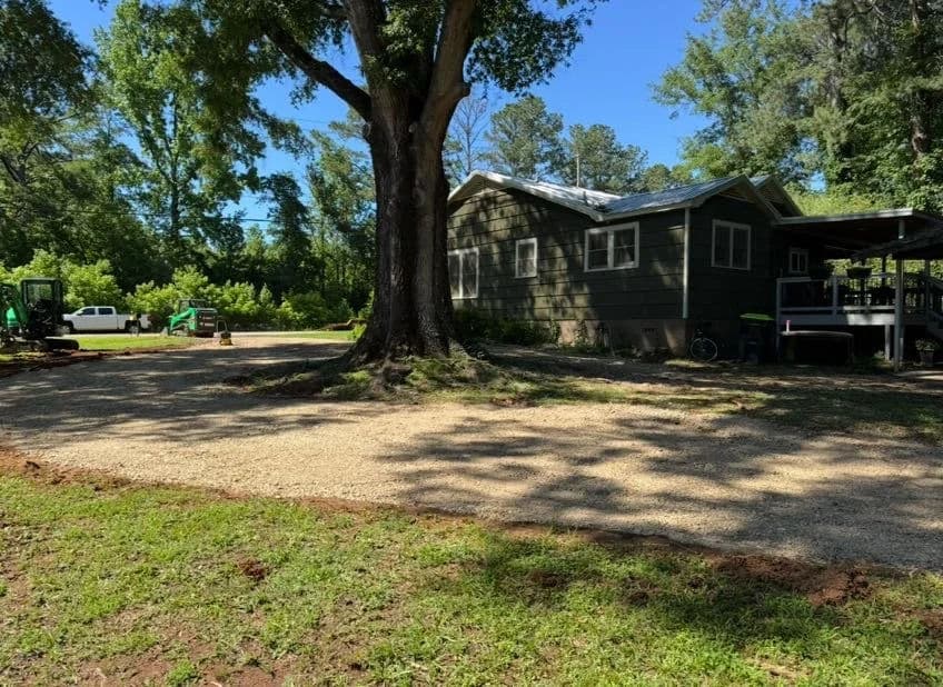 Green home surrounded by trees with a gravel driveway and equipment nearby.