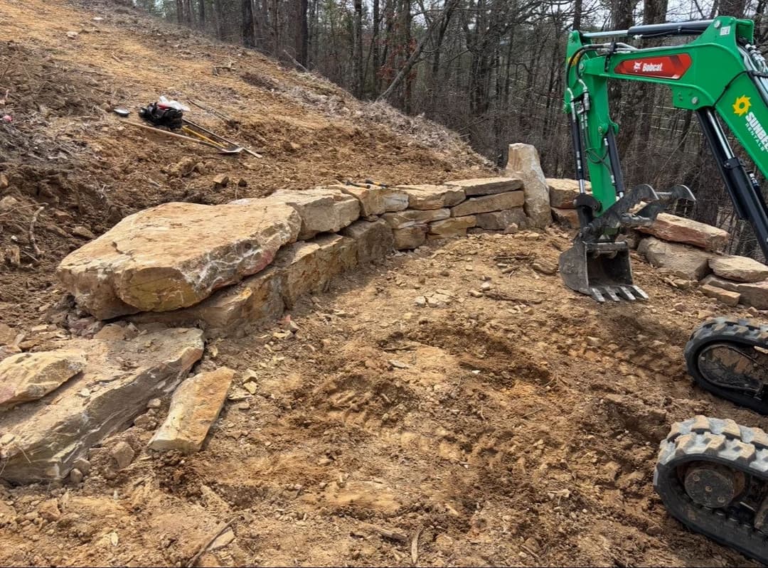 Excavator near a newly built stone retaining wall on a construction site in a forested area.