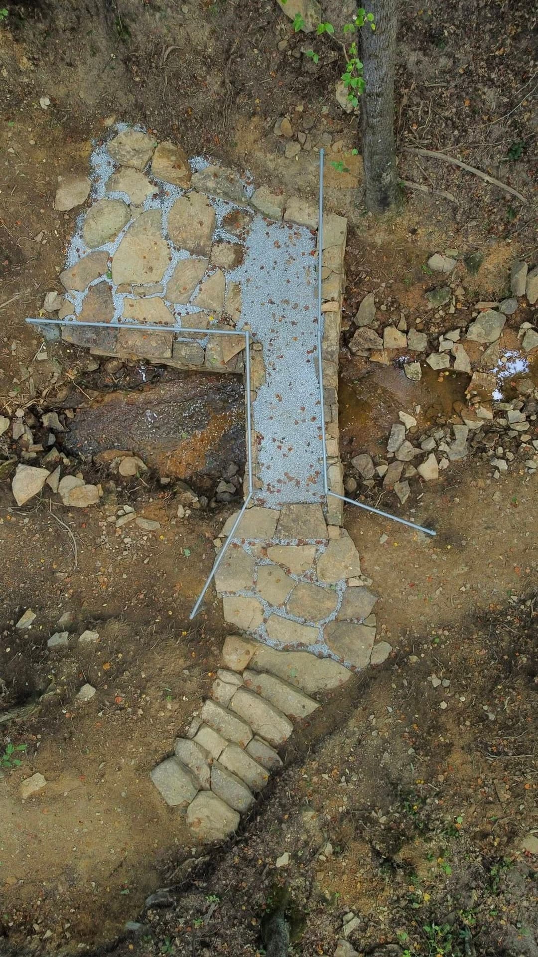 Aerial view of a stone pathway and bridge over a small stream in a wooded area.