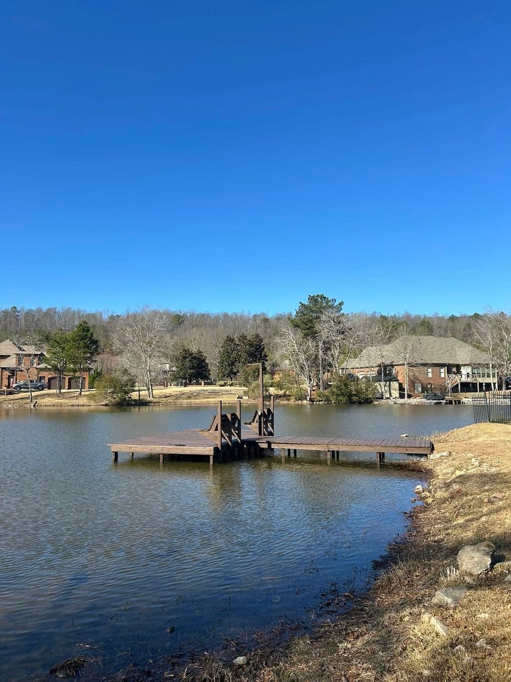Wooden dock over tranquil lake with clear blue sky and surrounding trees and houses.
