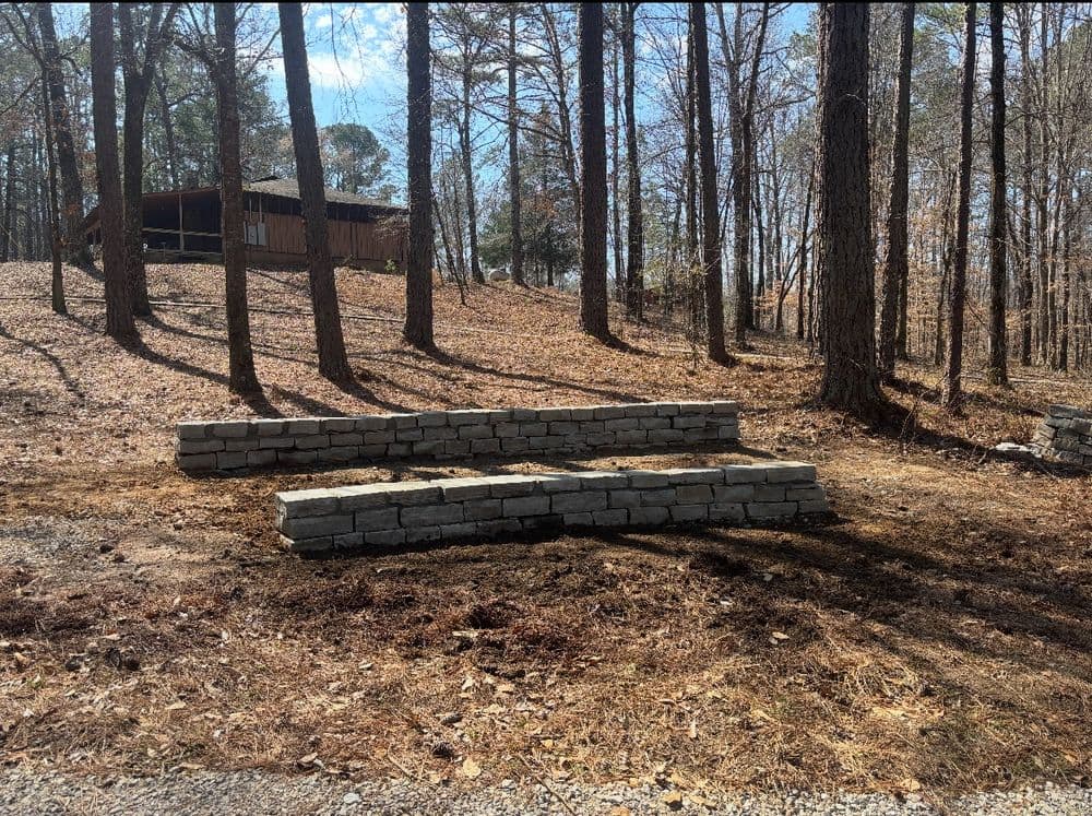 Stone retaining wall in a wooded area with a cabin in the background and fallen leaves.