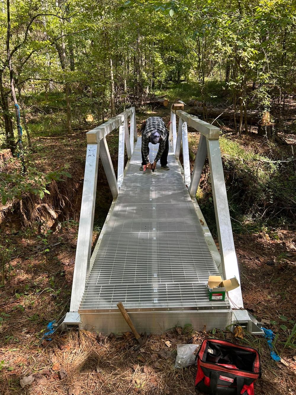 Worker assembling a metal bridge in a forested area, surrounded by greenery and tools.