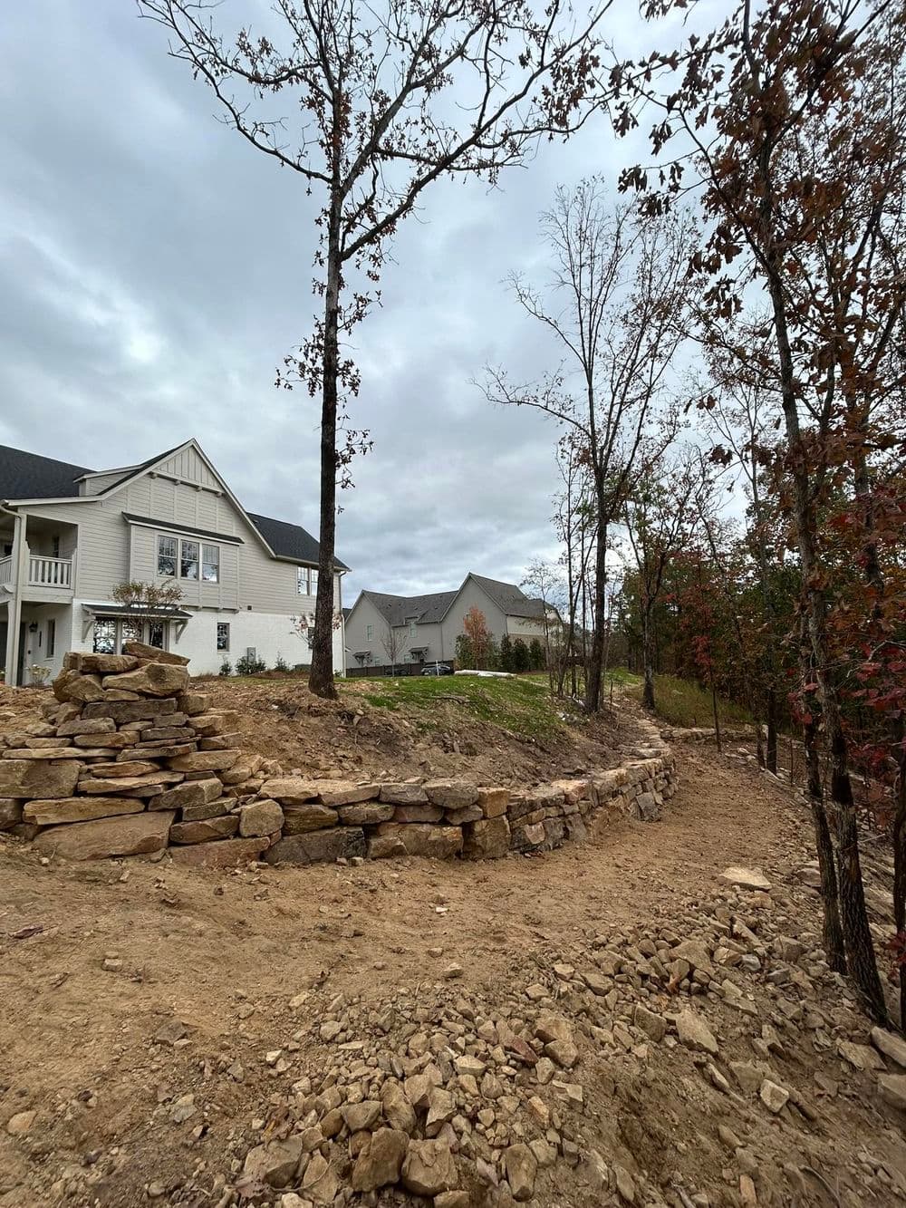 Newly landscaped property featuring stone wall, trees, and modern homes under cloudy sky.